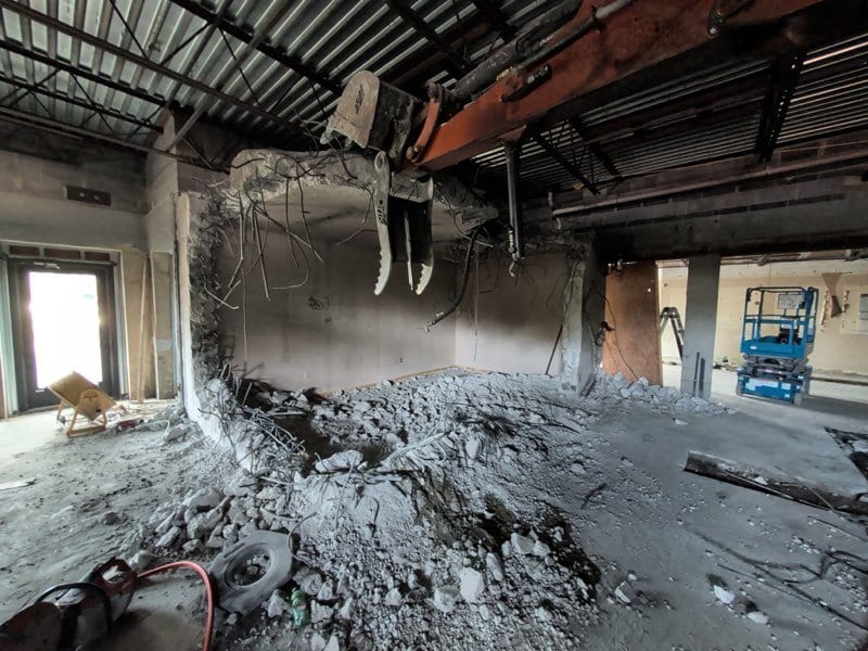 Demolition machine breaking out an interior wall, with concrete rubble piled on the floor